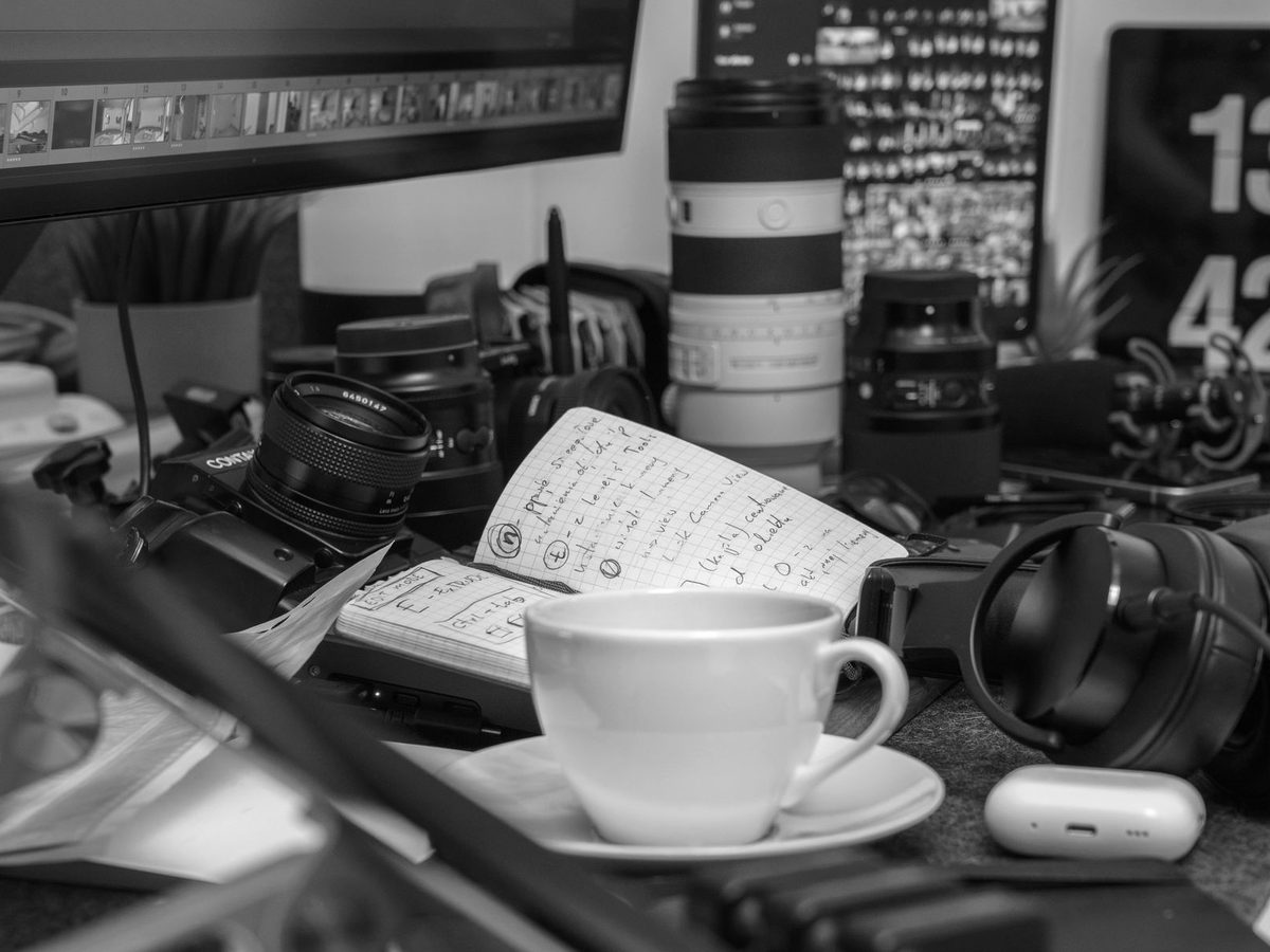 A working desk covered with camera lenses, headphones, notes and a coffee cup